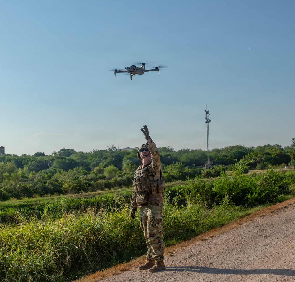 A military or law enforcement personnel in full camouflage uniform and tactical vest is launching or retrieving a quadcopter drone by hand on a gravel path. The operator is reaching upward toward the drone with one gloved hand while looking up at it. The setting appears to be a rural or semi-rural outdoor area with dense vegetation and a communications tower visible in the background.