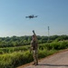 A military or law enforcement personnel in full camouflage uniform and tactical vest is launching or retrieving a quadcopter drone by hand on a gravel path. The operator is reaching upward toward the drone with one gloved hand while looking up at it. The setting appears to be a rural or semi-rural outdoor area with dense vegetation and a communications tower visible in the background.