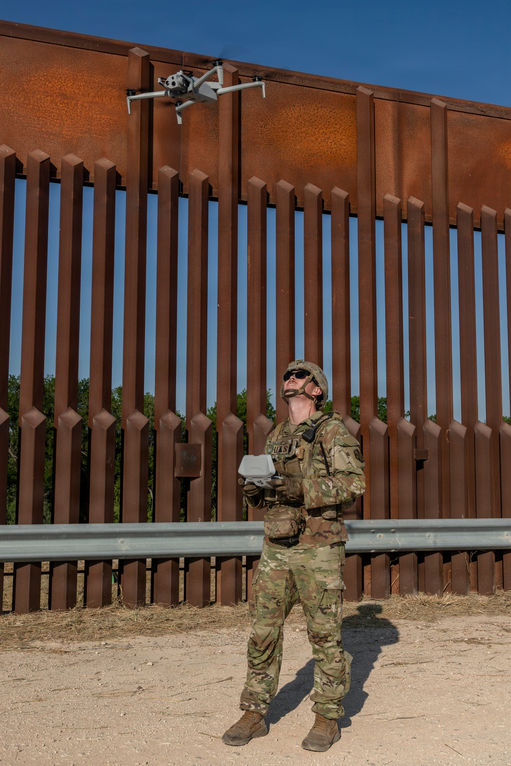 A uniformed military or law enforcement soldier in multicam/OCP camouflage is operating a small quadcopter drone near a large rust-colored steel border barrier. The operator is holding a remote controller and looking upward at the drone, which is hovering near the top of the barrier. The setting appears to be a dry, arid environment consistent with a U.S.-Mexico border region.