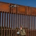 A uniformed military or law enforcement soldier in multicam/OCP camouflage is operating a small quadcopter drone near a large rust-colored steel border barrier. The operator is holding a remote controller and looking upward at the drone, which is hovering near the top of the barrier. The setting appears to be a dry, arid environment consistent with a U.S.-Mexico border region.