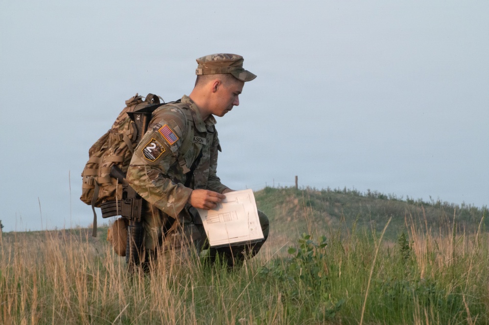 A U.S. Army soldier in full combat gear and OCP (Operational Camouflage Pattern) uniform is crouching in a grassy field while reviewing a map or document. The soldier is wearing a tactical backpack and appears to be conducting land navigation training. The setting appears to be an open training area with rolling terrain in the background.
