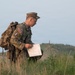 A U.S. Army soldier in full combat gear and OCP (Operational Camouflage Pattern) uniform is crouching in a grassy field while reviewing a map or document. The soldier is wearing a tactical backpack and appears to be conducting land navigation training. The setting appears to be an open training area with rolling terrain in the background.