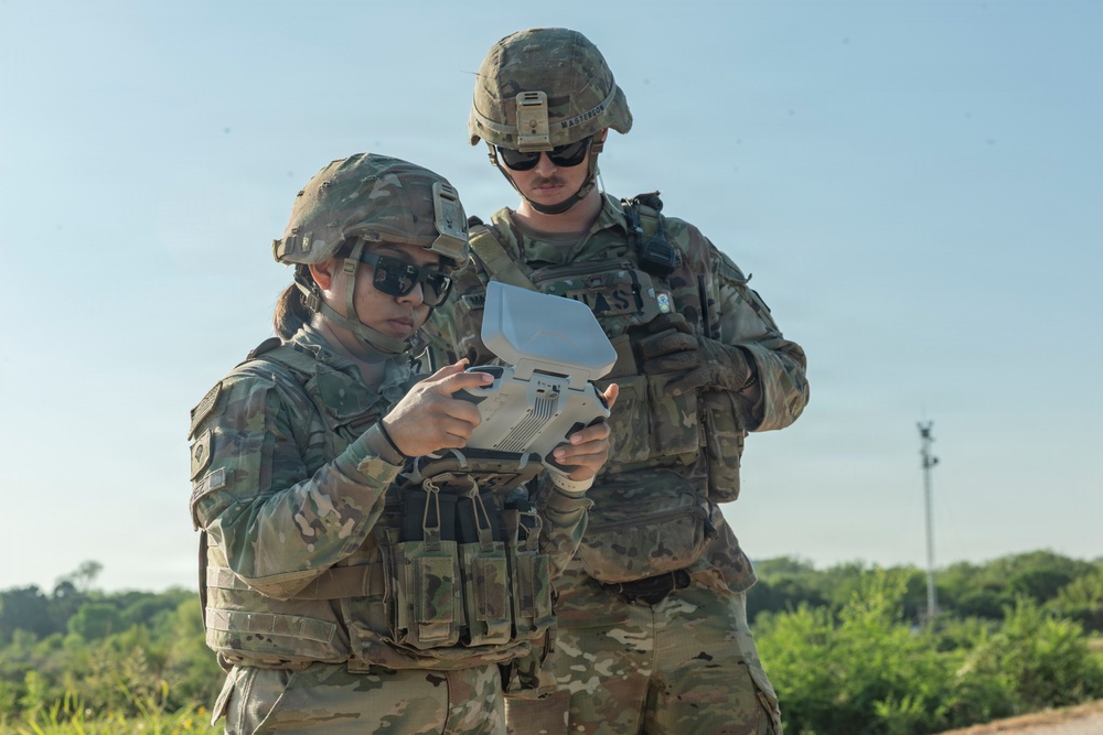 Two soldiers in U.S. Army OCP (Operational Camouflage Pattern) uniforms are operating what appears to be a drone ground control station or tactical handheld device outdoors. Both are wearing combat helmets, body armor, and sunglasses, suggesting a training or operational environment. The soldier in the foreground is actively manipulating the device while the second soldier observes.