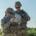 Two soldiers in U.S. Army OCP (Operational Camouflage Pattern) uniforms are operating what appears to be a drone ground control station or tactical handheld device outdoors. Both are wearing combat helmets, body armor, and sunglasses, suggesting a training or operational environment. The soldier in the foreground is actively manipulating the device while the second soldier observes.