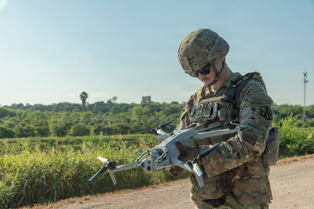 A U.S. Army soldier in full combat gear is holding and inspecting a small military-grade quadcopter drone. The soldier is wearing MultiCam (OCP) camouflage uniform, a tactical helmet with the name 'MASTERSON' visible, and sunglasses. The setting appears to be an outdoor training or operational environment with green vegetation and a clear sky in the background.