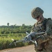 A U.S. Army soldier in full combat gear is holding and inspecting a small military-grade quadcopter drone. The soldier is wearing MultiCam (OCP) camouflage uniform, a tactical helmet with the name 'MASTERSON' visible, and sunglasses. The setting appears to be an outdoor training or operational environment with green vegetation and a clear sky in the background.