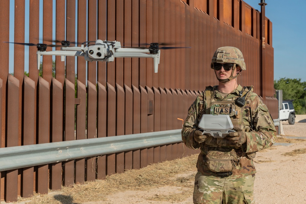 A uniformed military or law enforcement operator wearing OCP (Operational Camouflage Pattern) gear and a chest patch reading 'UAS' is piloting a quadcopter drone near a large rust-colored steel border barrier. The operator holds a remote controller and is observing the hovering drone. A vehicle is visible in the background to the right.