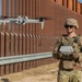 A uniformed military or law enforcement operator wearing OCP (Operational Camouflage Pattern) gear and a chest patch reading 'UAS' is piloting a quadcopter drone near a large rust-colored steel border barrier. The operator holds a remote controller and is observing the hovering drone. A vehicle is visible in the background to the right.