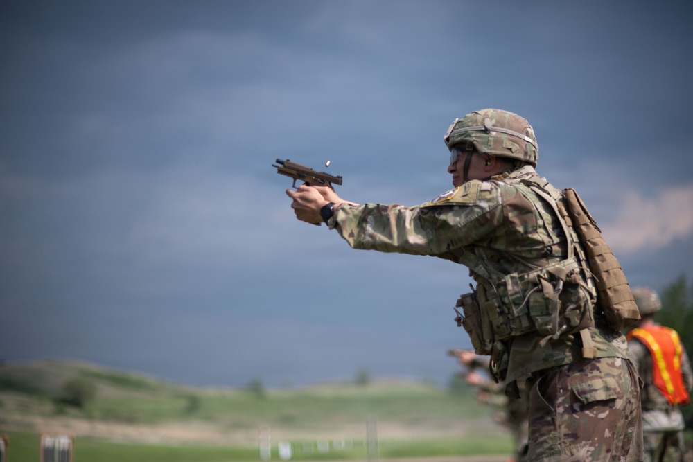 A soldier in full combat gear is firing a semi-automatic pistol at an outdoor shooting range. The soldier is wearing OCP (Operational Camouflage Pattern) uniform, helmet, body armor, and a backpack. At least one additional person in an orange safety vest is visible in the background, suggesting a supervised range exercise.