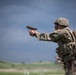 A soldier in full combat gear is firing a semi-automatic pistol at an outdoor shooting range. The soldier is wearing OCP (Operational Camouflage Pattern) uniform, helmet, body armor, and a backpack. At least one additional person in an orange safety vest is visible in the background, suggesting a supervised range exercise.