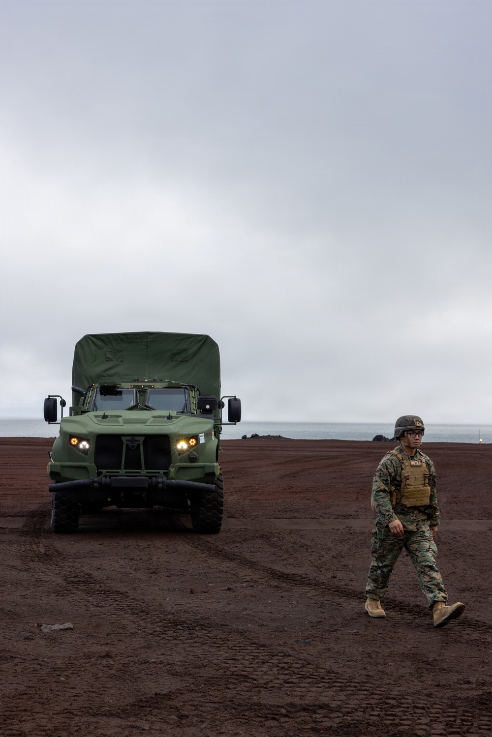 A uniformed military service member in camouflage battle dress walks across dark volcanic terrain near a coastline. Behind them, a green military medium tactical vehicle (MTV/MTVR) with headlights illuminated is parked or slowly moving on the same terrain. The overcast sky and dark reddish-brown ground suggest a coastal volcanic environment, possibly a Pacific island location.