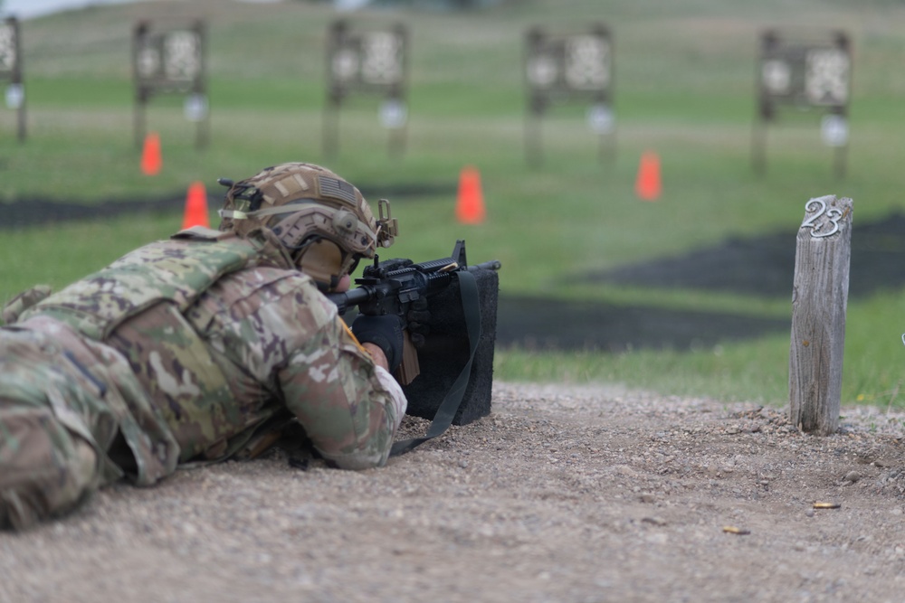 A soldier in full combat gear and MultiCam/OCP camouflage uniform is in the prone firing position on a military rifle range. The soldier is aiming a rifle, likely an M4 carbine, downrange toward multiple silhouette targets visible in the background. Orange safety cones and a numbered wooden post marked '23' indicate a designated firing lane.
