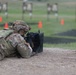 A soldier in full combat gear and MultiCam/OCP camouflage uniform is in the prone firing position on a military rifle range. The soldier is aiming a rifle, likely an M4 carbine, downrange toward multiple silhouette targets visible in the background. Orange safety cones and a numbered wooden post marked '23' indicate a designated firing lane.