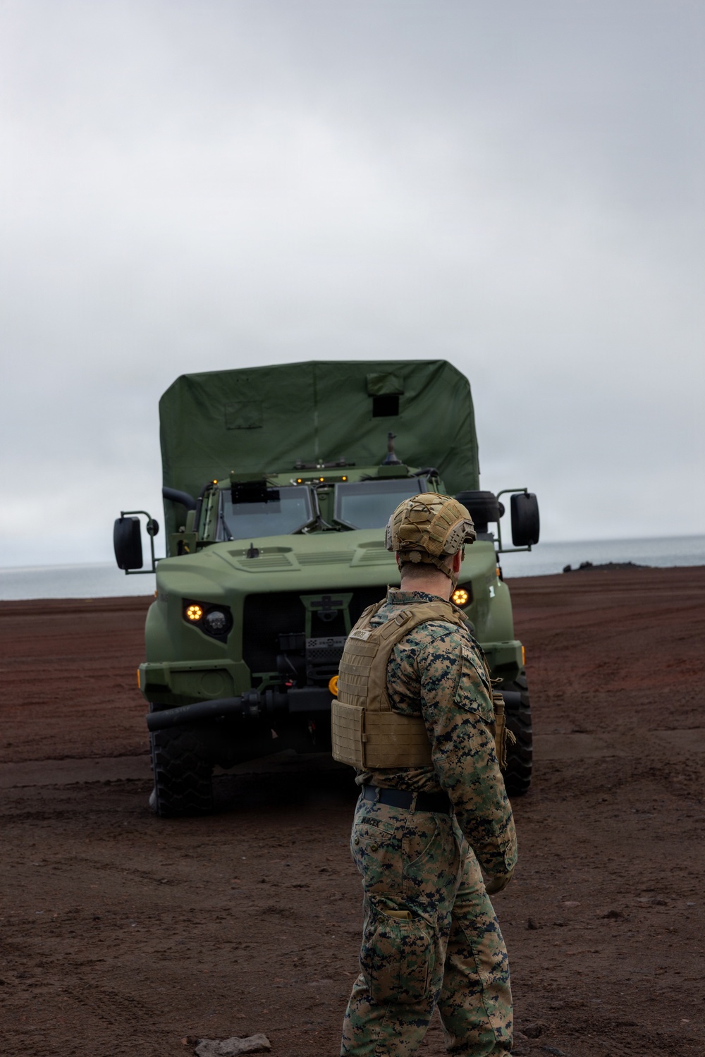 A military service member in digital camouflage (MARPAT) uniform and tactical gear stands facing a large olive-drab military truck on a dark, sandy coastal terrain under overcast skies. The vehicle appears to be an Oshkosh JLTV or similar modern military medium tactical vehicle with its headlights and marker lights illuminated. The setting suggests a beach landing or coastal operations exercise.