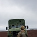 A military service member in digital camouflage (MARPAT) uniform and tactical gear stands facing a large olive-drab military truck on a dark, sandy coastal terrain under overcast skies. The vehicle appears to be an Oshkosh JLTV or similar modern military medium tactical vehicle with its headlights and marker lights illuminated. The setting suggests a beach landing or coastal operations exercise.