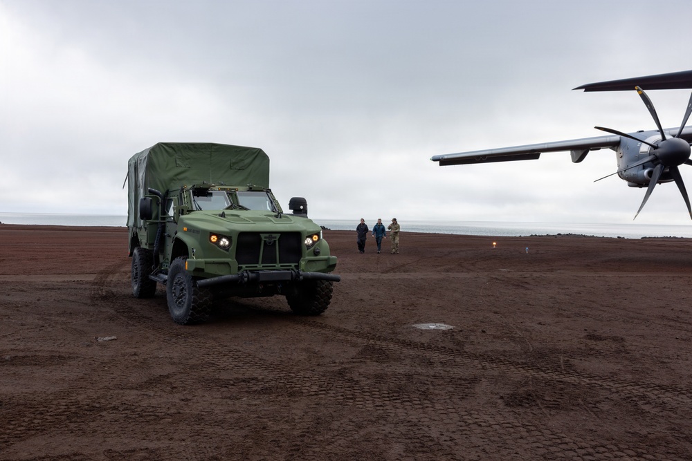 A military light tactical vehicle (appearing to be an JLTV or similar) is positioned on a dark, volcanic or tundra terrain near a propeller-driven aircraft. Three individuals are walking in the background toward the vehicle, with a large body of water visible on the horizon. The overcast sky and dark reddish-brown ground suggest a remote, possibly Arctic or sub-Arctic location.