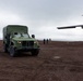 A military light tactical vehicle (appearing to be an JLTV or similar) is positioned on a dark, volcanic or tundra terrain near a propeller-driven aircraft. Three individuals are walking in the background toward the vehicle, with a large body of water visible on the horizon. The overcast sky and dark reddish-brown ground suggest a remote, possibly Arctic or sub-Arctic location.
