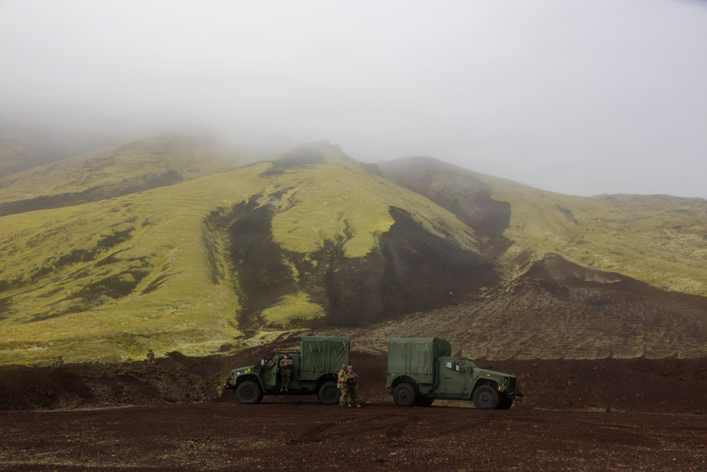 Two military tactical vehicles, consistent with JLTV or HMMWV variants, are parked on dark volcanic terrain in front of a moss-covered mountain partially obscured by low fog. Several personnel in military attire are visible near the vehicles. The landscape suggests a high-latitude volcanic environment, possibly Iceland or a similar region.