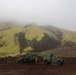 Two military tactical vehicles, consistent with JLTV or HMMWV variants, are parked on dark volcanic terrain in front of a moss-covered mountain partially obscured by low fog. Several personnel in military attire are visible near the vehicles. The landscape suggests a high-latitude volcanic environment, possibly Iceland or a similar region.