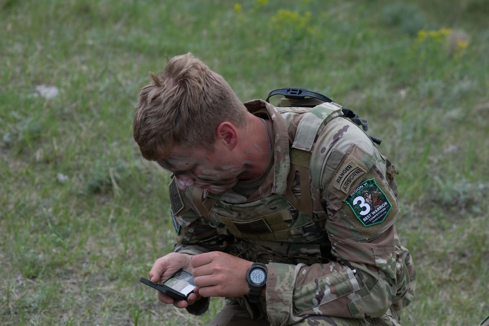 A soldier in MultiCam/OCP camouflage uniform is kneeling on grass while examining a small device, likely a compass or lensatic compass. The soldier has camouflage face paint applied and wears patches indicating Ranger and Airborne qualifications, along with a 'Best Warrior Competition' patch numbered '3' for what appears to be 'REGION VI'. The setting appears to be an outdoor field exercise or military competition event.