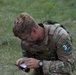 A soldier in MultiCam/OCP camouflage uniform is kneeling on grass while examining a small device, likely a compass or lensatic compass. The soldier has camouflage face paint applied and wears patches indicating Ranger and Airborne qualifications, along with a 'Best Warrior Competition' patch numbered '3' for what appears to be 'REGION VI'. The setting appears to be an outdoor field exercise or military competition event.
