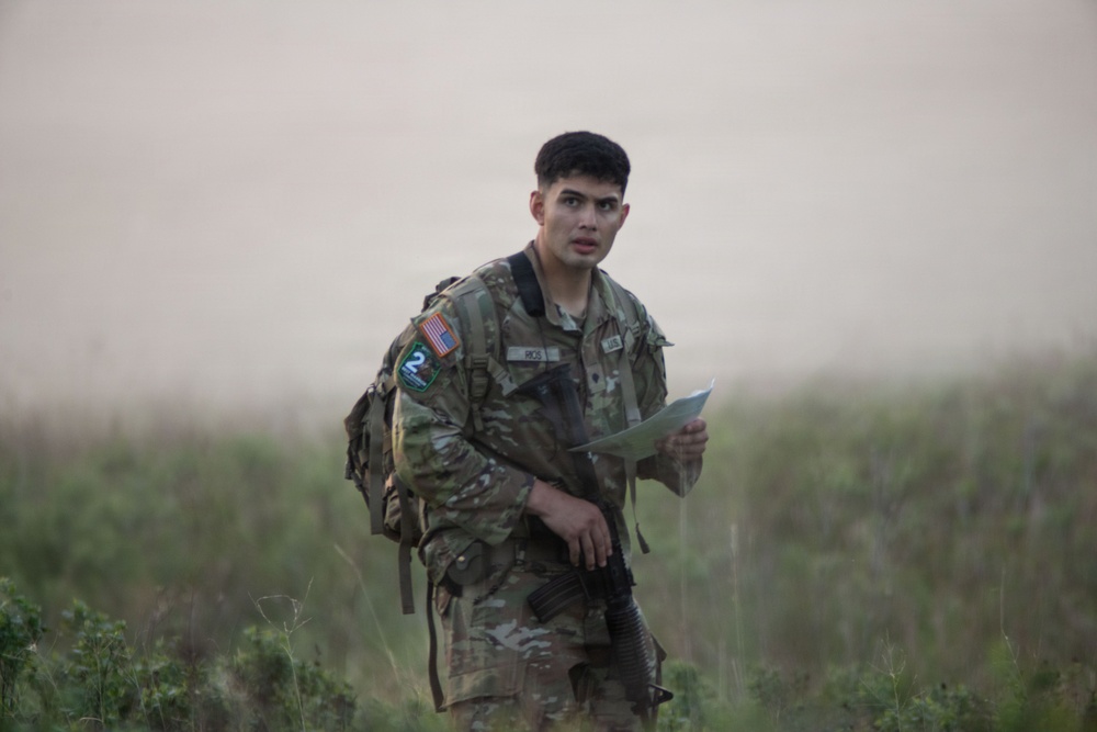A U.S. Army soldier in MultiCam/OCP camouflage uniform is standing in an open grassy field, holding what appears to be a map or document. He is equipped with a rucksack and carries a rifle, suggesting he is engaged in a land navigation or field training exercise. The background is hazy and out of focus, with green vegetation visible in the foreground.
