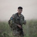 A U.S. Army soldier in MultiCam/OCP camouflage uniform is standing in an open grassy field, holding what appears to be a map or document. He is equipped with a rucksack and carries a rifle, suggesting he is engaged in a land navigation or field training exercise. The background is hazy and out of focus, with green vegetation visible in the foreground.