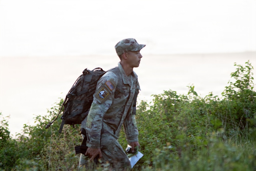 A U.S. Army soldier in ACU (Army Combat Uniform) camouflage is navigating through dense vegetation near what appears to be a body of water in the background. The soldier is carrying a military rucksack and holding what appears to be a map or document, suggesting a land navigation exercise or field training operation. The image is taken in natural outdoor lighting with a hazy, overcast sky.