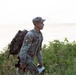 A U.S. Army soldier in ACU (Army Combat Uniform) camouflage is navigating through dense vegetation near what appears to be a body of water in the background. The soldier is carrying a military rucksack and holding what appears to be a map or document, suggesting a land navigation exercise or field training operation. The image is taken in natural outdoor lighting with a hazy, overcast sky.