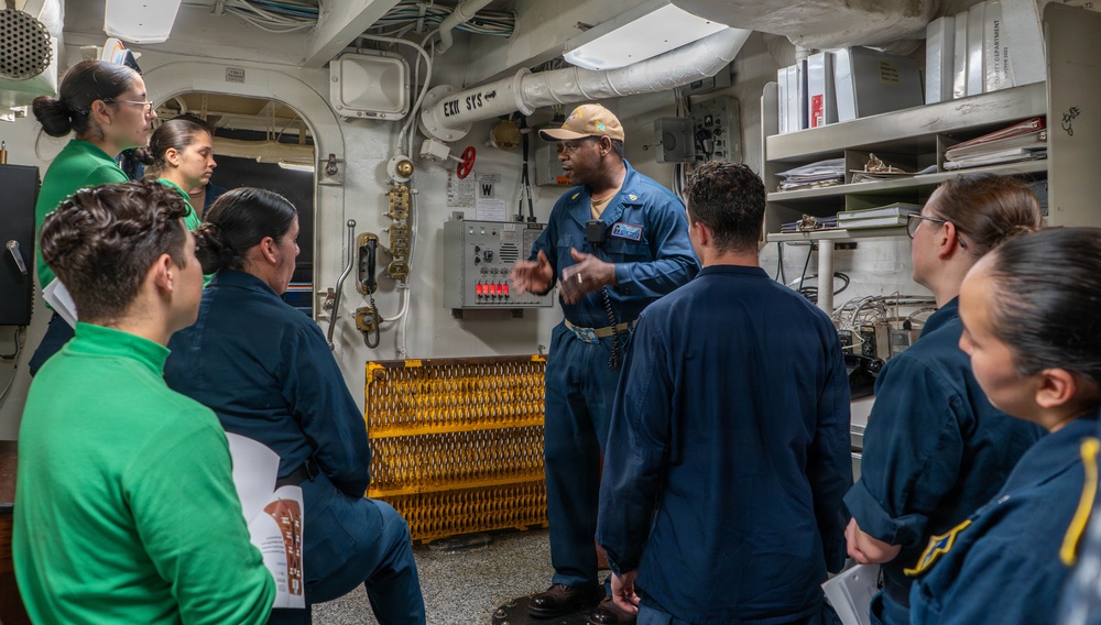 A military instructor or senior enlisted personnel in blue coveralls and a cap is conducting a briefing or training session aboard what appears to be a U.S. Navy vessel. A group of approximately seven personnel, some in blue coveralls and two in green athletic wear, are gathered in a shipboard compartment listening attentively. The setting includes visible ship infrastructure such as piping, a control panel, wall-mounted telephone, and storage shelves with binders.