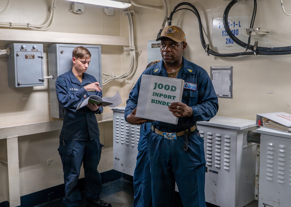 Two U.S. Navy personnel are shown inside what appears to be a shipboard compartment or watch station. The individual in the foreground is holding a binder labeled 'JOOD INPORT BINDER,' while a second sailor in the background reviews documents. The setting is consistent with an in-port watch duty station aboard a U.S. Navy vessel.