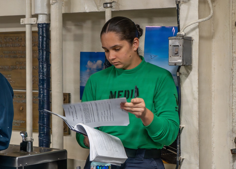 A young woman wearing a green sweatshirt with 'MEDIA' printed on it is reading through a multi-page document or booklet. She appears to be standing in an interior space that resembles a naval vessel, evidenced by the bronze dedication plaque partially visible on the left wall referencing 'George W...' and 'nuclear-powered aircraft.' The setting includes industrial-style walls, pipes, and a photograph of aircraft in the background.