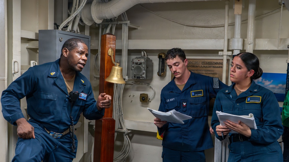 Three U.S. Navy personnel are engaged in what appears to be a briefing or training session aboard a naval vessel, identified by a plaque as the USS George Washington nuclear-powered aircraft carrier. A senior sailor on the left is speaking and gesturing while two junior sailors hold and review documents. The setting is an interior shipboard compartment featuring a brass ship's bell and various pipes and cables.
