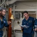 Three U.S. Navy personnel are engaged in what appears to be a briefing or training session aboard a naval vessel, identified by a plaque as the USS George Washington nuclear-powered aircraft carrier. A senior sailor on the left is speaking and gesturing while two junior sailors hold and review documents. The setting is an interior shipboard compartment featuring a brass ship's bell and various pipes and cables.