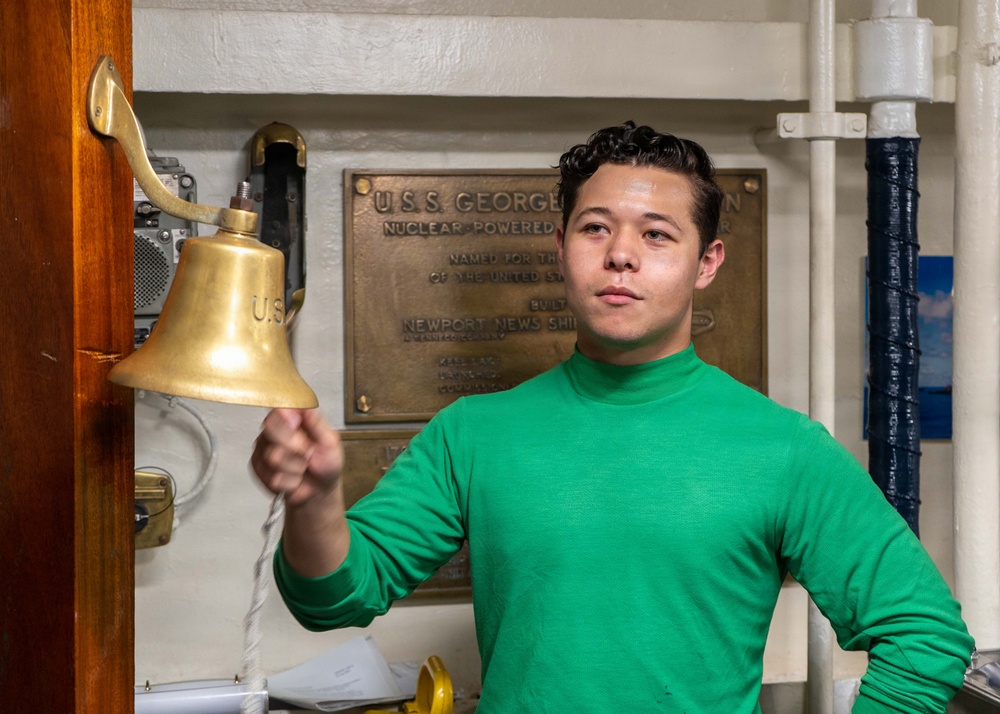 A young man wearing a green long-sleeve turtleneck shirt is holding a brass ship's bell by its rope lanyard. He is standing in front of a bronze dedication plaque that reads 'U.S.S. George [Washington]' and references 'Nuclear Powered' and 'Newport News Shipbuilding.' The setting appears to be aboard a naval vessel, likely a museum ship or active carrier.