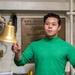 A young man wearing a green long-sleeve turtleneck shirt is holding a brass ship's bell by its rope lanyard. He is standing in front of a bronze dedication plaque that reads 'U.S.S. George [Washington]' and references 'Nuclear Powered' and 'Newport News Shipbuilding.' The setting appears to be aboard a naval vessel, likely a museum ship or active carrier.