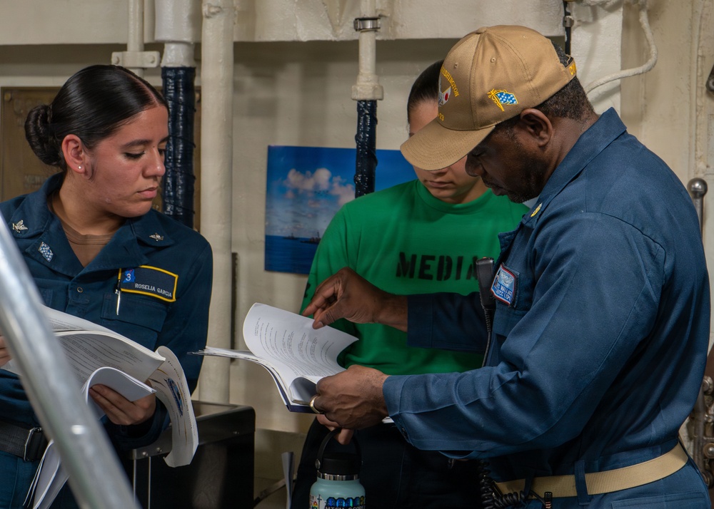 Three individuals are gathered in what appears to be a naval vessel interior, reviewing documents together. A female sailor in blue uniform with a name tag reading 'ROSELIA GARCIA' holds papers, while a senior male sailor in blue coveralls and a tan cap examines a binder or booklet. A third person in a green 'MEDIA' shirt stands in the background observing.