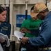 Three individuals are gathered in what appears to be a naval vessel interior, reviewing documents together. A female sailor in blue uniform with a name tag reading 'ROSELIA GARCIA' holds papers, while a senior male sailor in blue coveralls and a tan cap examines a binder or booklet. A third person in a green 'MEDIA' shirt stands in the background observing.