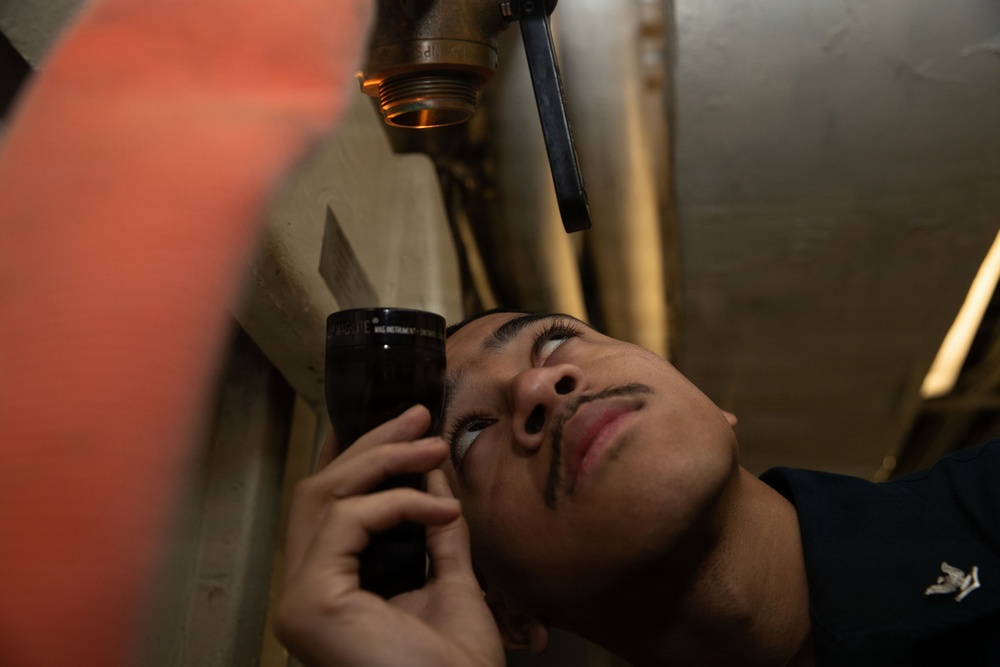 A person is using a black flashlight to inspect piping or valve components in a confined, dimly lit space. The individual is looking upward closely at a brass or bronze threaded valve/fitting, suggesting a maintenance or inspection task. The environment appears to be an industrial or mechanical space such as a ship's interior or utility room.