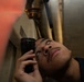 A person is using a black flashlight to inspect piping or valve components in a confined, dimly lit space. The individual is looking upward closely at a brass or bronze threaded valve/fitting, suggesting a maintenance or inspection task. The environment appears to be an industrial or mechanical space such as a ship's interior or utility room.
