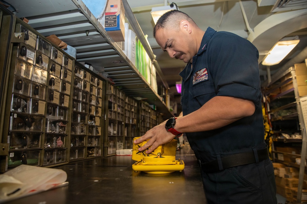 A military personnel member in a dark navy uniform is working in what appears to be a shipboard supply or parts storage room. He is handling a bright yellow piece of equipment or component on a workbench. The room is lined with numerous small parts drawers and shelving units typical of a military supply depot or ship's storeroom.