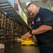 A military personnel member in a dark navy uniform is working in what appears to be a shipboard supply or parts storage room. He is handling a bright yellow piece of equipment or component on a workbench. The room is lined with numerous small parts drawers and shelving units typical of a military supply depot or ship's storeroom.