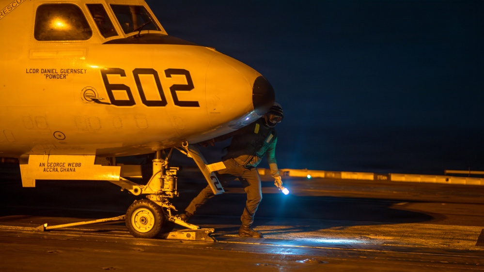A nighttime scene on what appears to be an aircraft carrier flight deck, showing a military aircraft numbered '602' with the name 'LCDR DANIEL GUERNSEY POWDER' stenciled on the fuselage. A deck crew member in a green jersey is crouching near the nose landing gear, holding a light wand. The aircraft also bears the text 'AN GEORGE WEBB ACCRA, GHANA' near the nose gear area.