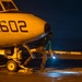 A nighttime scene on what appears to be an aircraft carrier flight deck, showing a military aircraft numbered '602' with the name 'LCDR DANIEL GUERNSEY POWDER' stenciled on the fuselage. A deck crew member in a green jersey is crouching near the nose landing gear, holding a light wand. The aircraft also bears the text 'AN GEORGE WEBB ACCRA, GHANA' near the nose gear area.