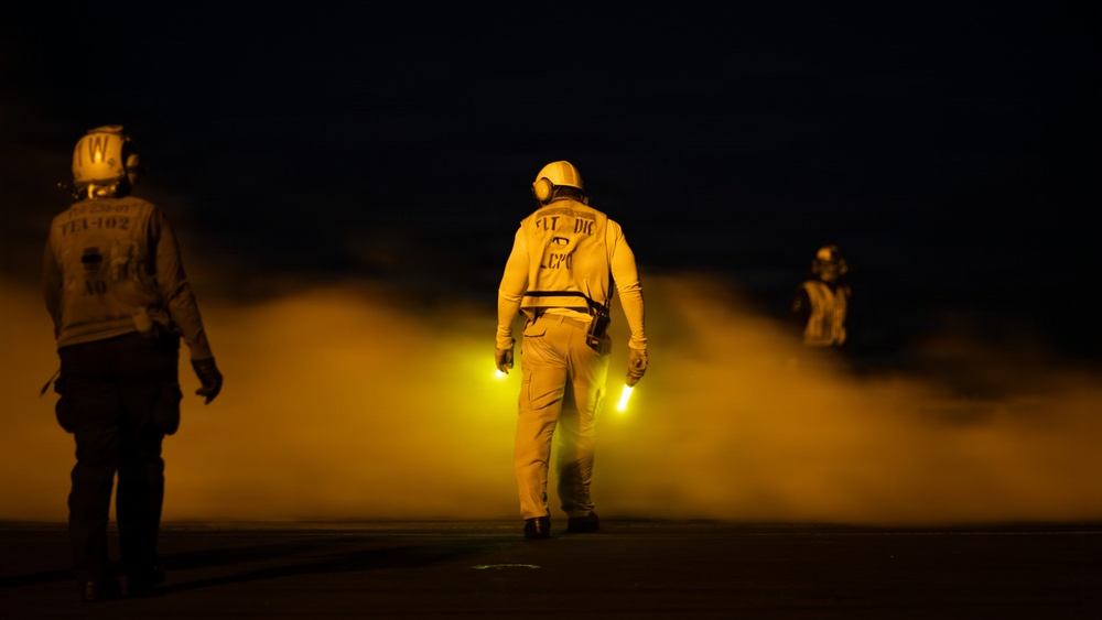 A nighttime scene on what appears to be an aircraft carrier flight deck, with personnel in yellow flight deck jerseys and helmets moving through steam or exhaust vapor. The central figure holds illuminated yellow glow sticks used for signaling, while two other crew members are visible in the background. The dramatic lighting from the glow sticks and ambient yellow light creates a high-contrast, atmospheric image.