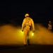A nighttime scene on what appears to be an aircraft carrier flight deck, with personnel in yellow flight deck jerseys and helmets moving through steam or exhaust vapor. The central figure holds illuminated yellow glow sticks used for signaling, while two other crew members are visible in the background. The dramatic lighting from the glow sticks and ambient yellow light creates a high-contrast, atmospheric image.