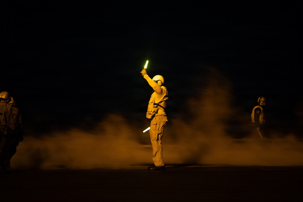 A night scene showing a person in flight deck gear holding illuminated glow sticks aloft, likely directing aircraft on a naval carrier flight deck. The figure is surrounded by exhaust or steam clouds, with at least two other similarly equipped personnel visible in the background. The scene is characteristic of nighttime aircraft carrier flight deck operations.