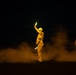 A night scene showing a person in flight deck gear holding illuminated glow sticks aloft, likely directing aircraft on a naval carrier flight deck. The figure is surrounded by exhaust or steam clouds, with at least two other similarly equipped personnel visible in the background. The scene is characteristic of nighttime aircraft carrier flight deck operations.