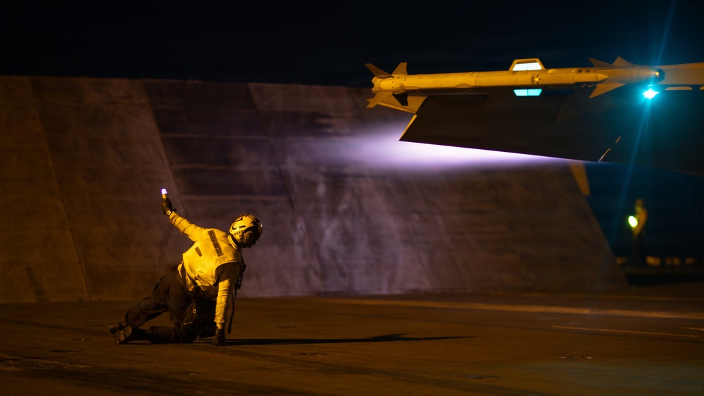 A night operations scene on what appears to be an aircraft carrier flight deck, showing a deck crew member in a crouching position holding a illuminated wand. A military aircraft wing is visible overhead, equipped with what appears to be an air-to-air missile (likely an AIM-9 Sidewinder) on a wingtip rail. The scene is dramatically lit by the aircraft's landing/taxi light and navigation lights.