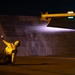 A night operations scene on what appears to be an aircraft carrier flight deck, showing a deck crew member in a crouching position holding a illuminated wand. A military aircraft wing is visible overhead, equipped with what appears to be an air-to-air missile (likely an AIM-9 Sidewinder) on a wingtip rail. The scene is dramatically lit by the aircraft's landing/taxi light and navigation lights.